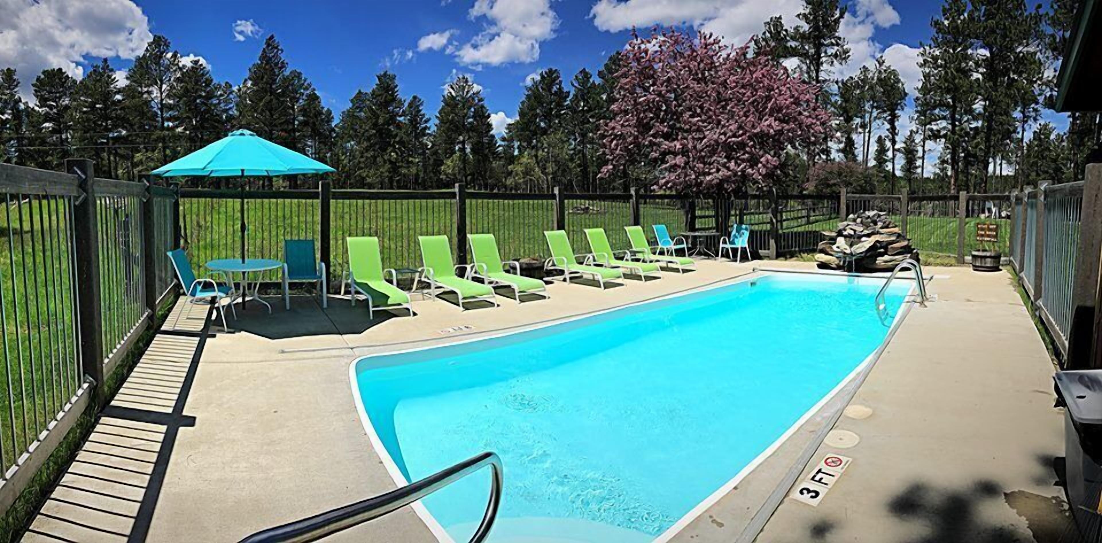 Piscina rectangular con cubierta de madera, rodeada por una cerca y sillas reclinables verdes.