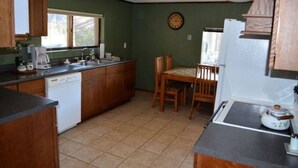 A compact kitchen with wooden cabinets, a dishwasher, a dining table with chairs, and a clock on the wall.
