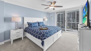 A bedroom with a bed, nightstands, a ceiling fan, a TV, and a large window with a view of a building. - Beachfront Condo | Views, Pool & Tennis Courts (Marco Island)