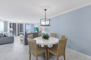 A dining area with a round table, wooden chairs, and a hanging light fixture. - Beachfront Condo | Views, Pool & Tennis Courts (Marco Island)