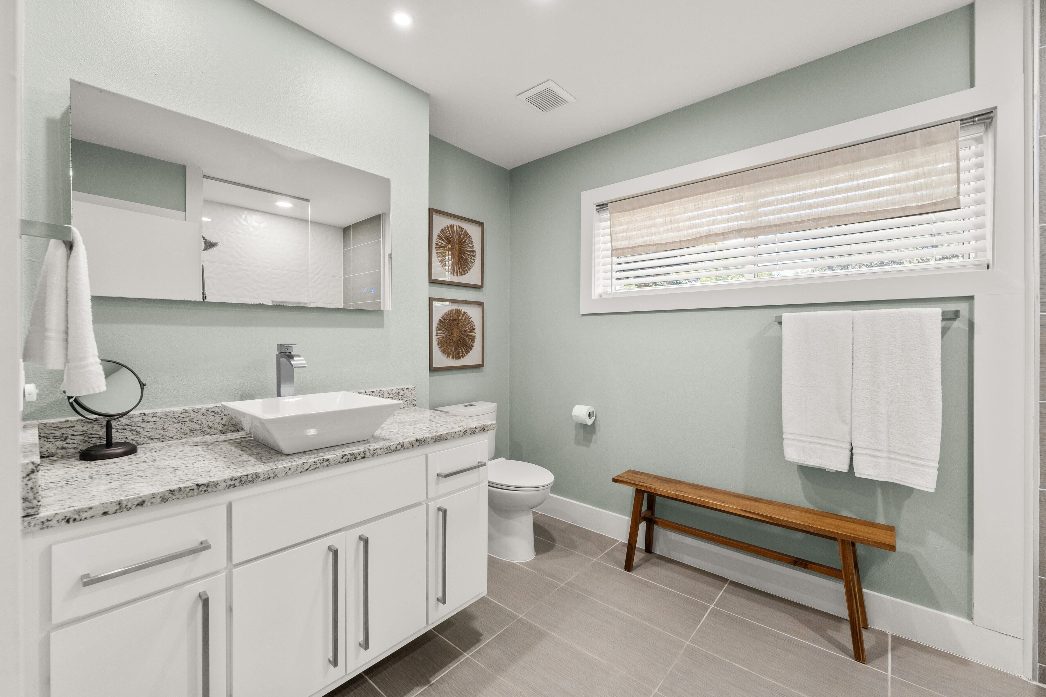 Ein modernes Badezimmer mit Waschbecken, Waschtisch und Toilette. Eine Sitzbank und ein Fenster mit Jalousien sind ebenfalls vorhanden.