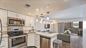 A modern kitchen with stainless steel appliances, a dishwasher, and a built-in oven. There is a large island with a sink and a seating area with a sofa and cushions. The kitchen has a wooden floor and white cabinetry.