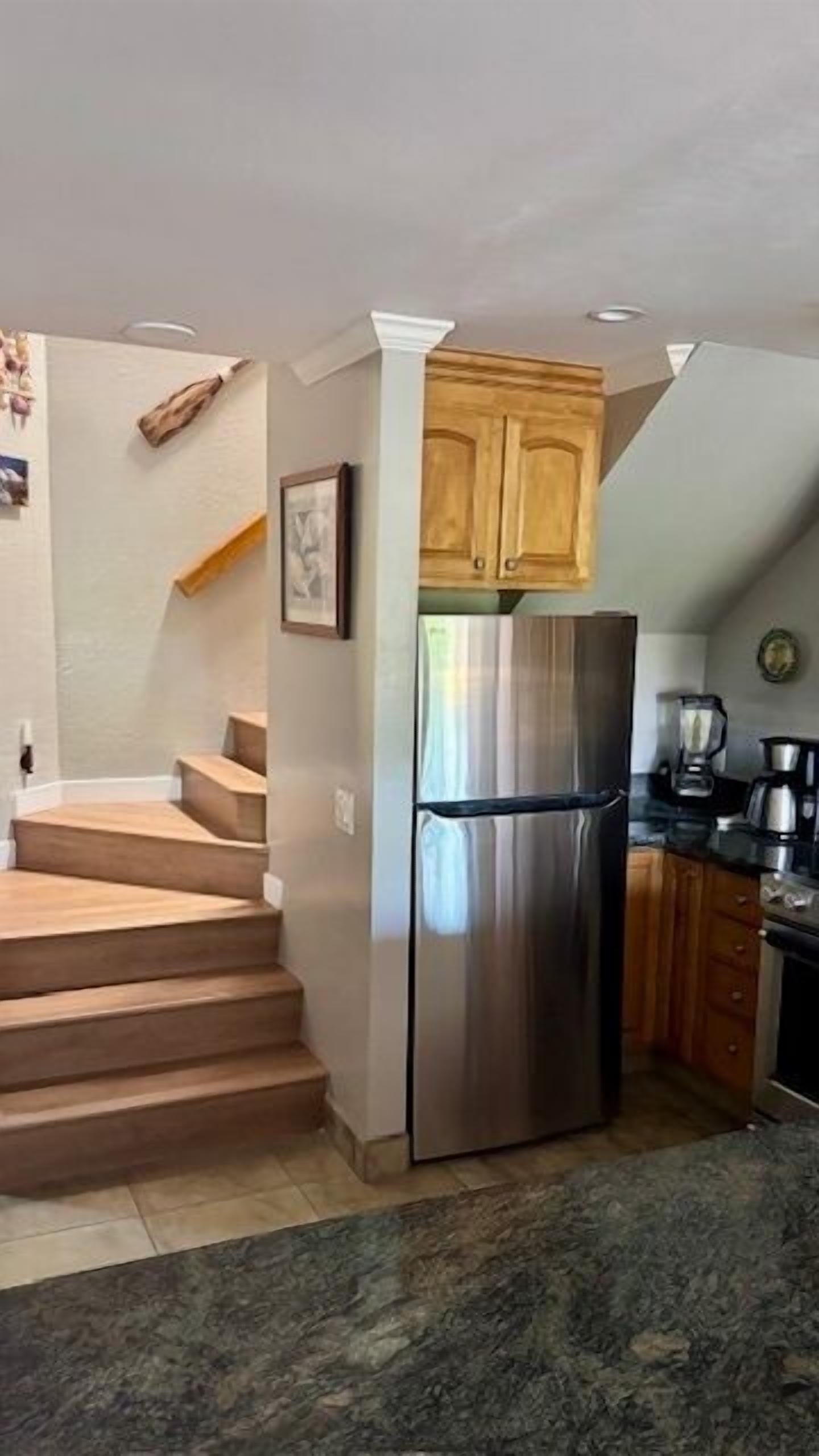 Una cocina con refrigerador de acero inoxidable, gabinetes de madera, cafetera y una escalera que lleva a otro nivel.