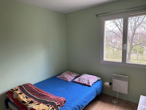 A single bed with a blue mattress, two pillows, a patterned blanket, a window with a view of trees, and a wall-mounted heater.