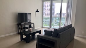 A living room with a flat-screen TV on a stand, a dark coffee table, a gray sofa, and a floor lamp. - Beach Front Spacious 1BR , The Tides, Hollywood,FL (Hollywood)