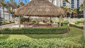 A thatched-roof outdoor seating area with tables and chairs, surrounded by well-maintained hedges and palm trees, with a white fence and high-rise buildings in the background. - Beach Front Spacious 1BR , The Tides, Hollywood,FL (Hollywood)