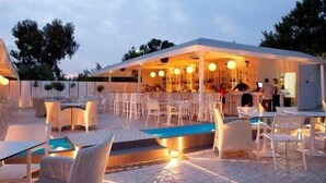 Un espace extérieur aménagé pour les repas, avec des tables et des chaises blanches, une piscine et un coin salon couvert agrémenté de guirlandes lumineuses.