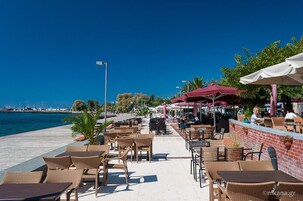 Café al aire libre con mobiliario de mimbre y sombrillas, con vista a un cuerpo de agua.