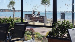 Vue depuis l’intérieur d’un bâtiment doté de grandes fenêtres donnant sur une plage. On aperçoit des chaises et des tables au premier plan, et un banc où sont assis deux personnes, à l’arrière-plan.