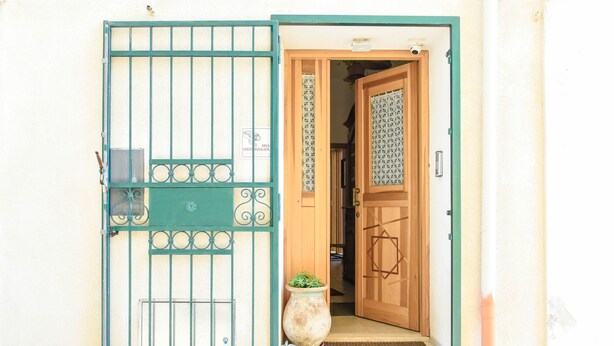 A building entrance with a green metal gate, a wooden door with a geometric pattern, and a potted plant on the steps. - Casa Marta (Nuoro)