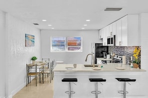 A modern kitchen with white cabinets, a central island with stools, a dining area with chairs, and a window with blinds.