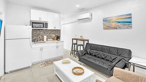 A modern living space with a kitchen, a black leather sofa, a small dining table, and a wall-mounted air conditioner.
