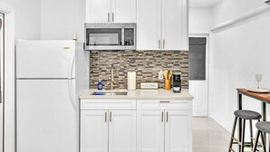 A modern kitchen with white cabinets, a stainless steel microwave, a tiled backsplash, and a small bar area with stools.