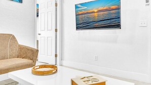 A room with a white wall, a framed picture of a sunset over the ocean, a beige sofa, and a white table with a tray and a book.