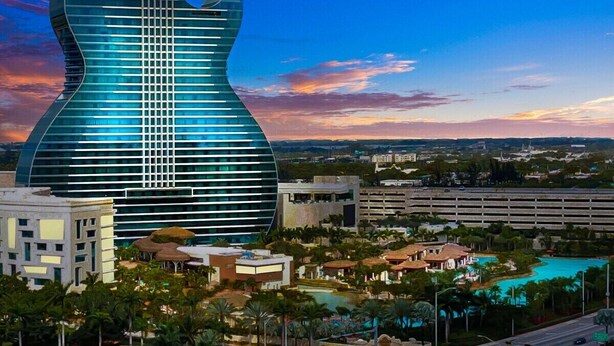 A modern building with a unique guitar-shaped design, surrounded by a resort area with pools and greenery.