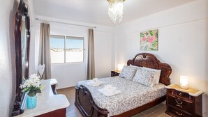 Ein Schlafzimmer mit einem großen Bett, einem Nachttisch, einem Spiegel, einem Fenster mit Vorhängen und einer Kristallleuchtre.