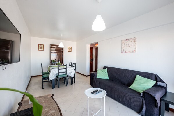 Ein Wohnzimmer mit einer Couch, einem Esstisch mit Stühlen, einem kleinen weißen Tisch und einem Fernseher an der Wand.