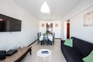 Un salon avec un canapé noir, une table basse blanche, un téléviseur à écran plat fixé au mur et un coin repas comprenant une table et des chaises.