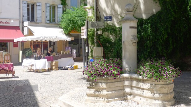 Una calle adoquinada con un parterre de flores, un café con terraza y un edificio con toldo rojo.