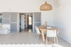 Uma zona de refeições moderna com mesa de madeira e cadeiras brancas, uma luminária pendente trançada e vista para o quarto adjacente.