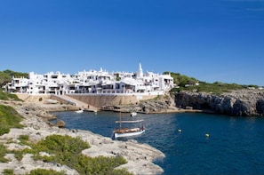 Uma vila costeira com edifícios brancos, costa rochosa e um pequeno barco ancorado perto da praia.