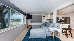 A modern living room with a large window, a gray sectional sofa, a white side table, and a kitchen area visible through an open door. - Serene Lakefront home in Tampa Bay, close to beaches and downtown Tarpon Springs (holiday)