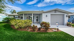 A modern single-story house with a covered porch, a garage, and a well-maintained front yard with various shrubs and a small garden. - Serene Lakefront home in Tampa Bay, close to beaches and downtown Tarpon Springs (holiday)