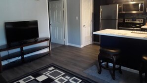 A compact kitchen with a refrigerator, microwave, and oven. There is a small island with stools, a television on a stand, and a patterned rug on the floor.