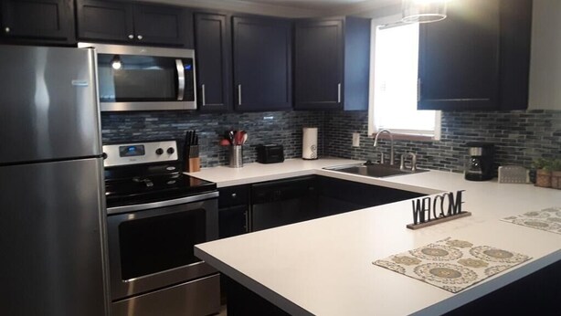 A modern kitchen with dark cabinets, stainless steel appliances, a large island, and a welcome mat.