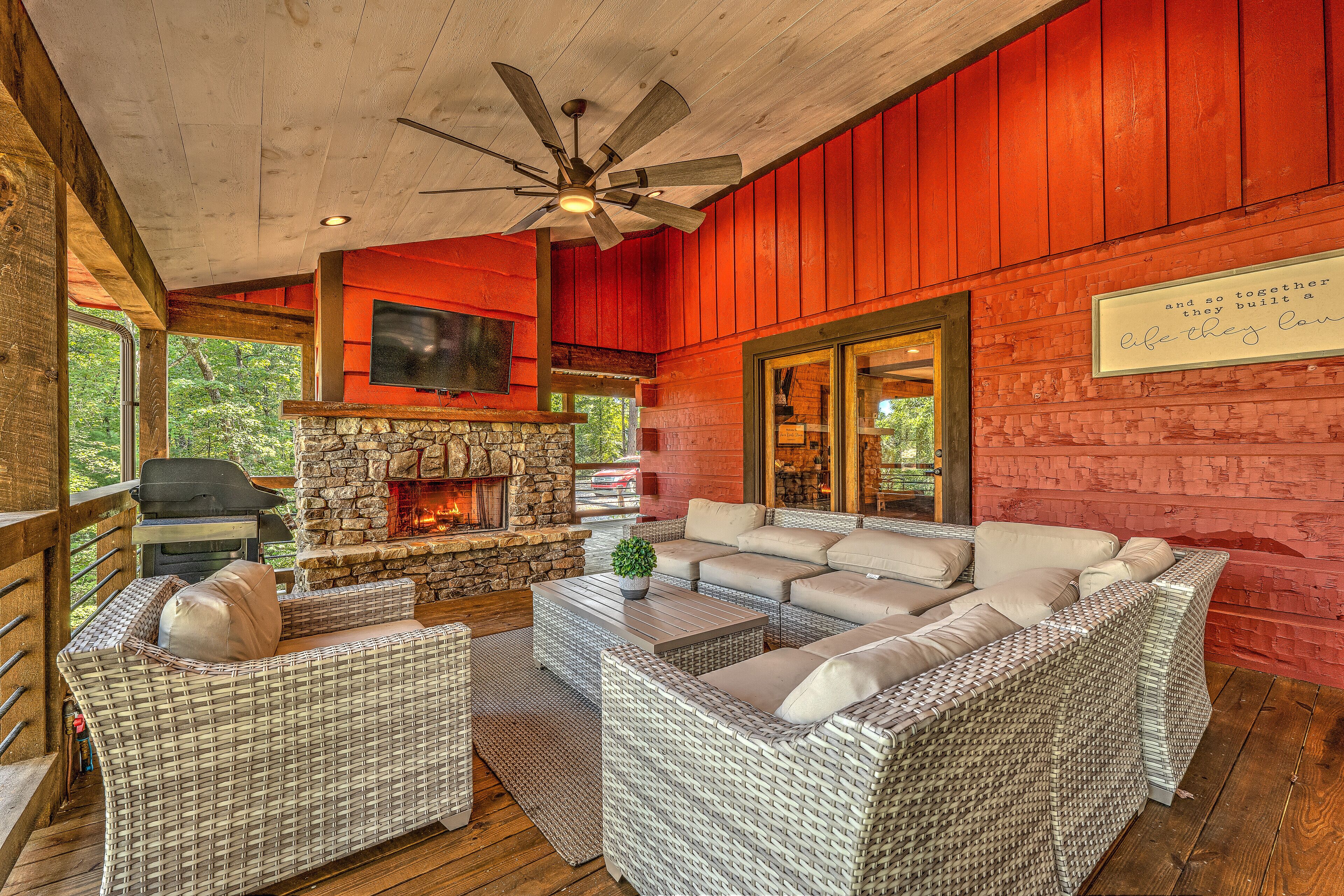 A cozy outdoor seating area with wicker furniture, a stone fireplace, and a ceiling fan.