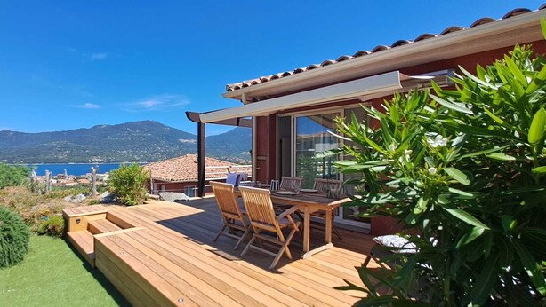 Un terrazzo in legno con tavolo e sedie, affacciato su un paesaggio costiero con montagne.