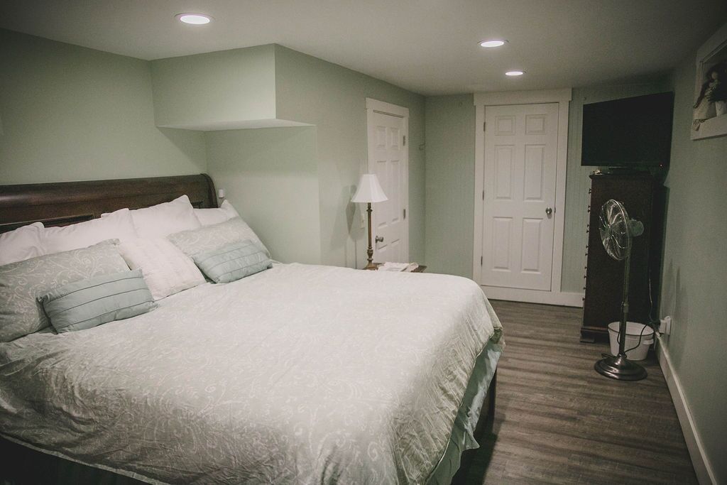 A neatly made bed with a patterned bedspread, a bedside lamp, and a standing fan in a room with light green walls and wooden flooring.