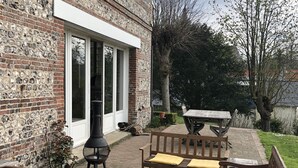 Een terras buiten met houten banken en een tafel, een bakstenen muur en een groot raam.