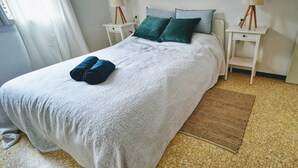 A neatly made bed with a white quilt, two pillows, and two rolled towels. There are two bedside tables with lamps, a small rug, and a window with curtains. - Beau T3 -terrasse & Parking, Proche Plage 1er Étage Droite (Hyères)
