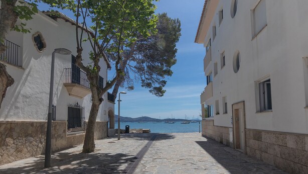 A cobblestone street flanked by white buildings with balconies, leading to a body of water with boats. - Apartamento Coral, Holiday Apartment in Puerto Pollença (Puerto Pollença)