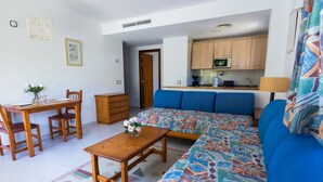 A living room with a sofa, a dining table with chairs, a wooden cabinet, and a kitchen area with a microwave. - Apartamento Coral, Holiday Apartment in Puerto Pollença (Puerto Pollença)