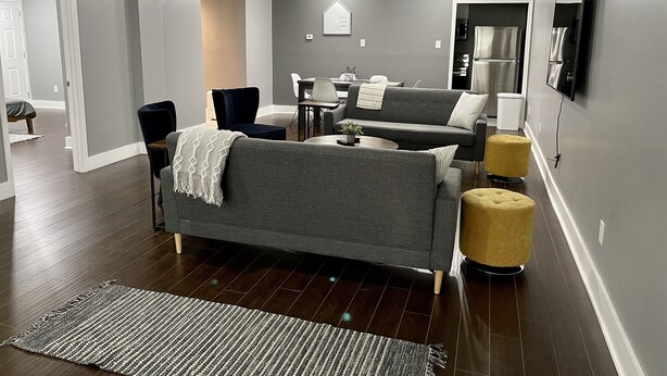Een moderne woonkamer met een grijze bank, een klein tafeltje en een geel poefje. Op de achtergrond is een eethoek met een tafel en stoelen te zien.