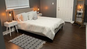 Een slaapkamer met een bed, twee nachtkastjes, een spiegel, een deur en een raam met jaloezieën.
