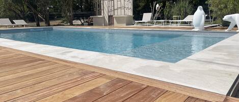 Uma piscina com deck de madeira, rodeada por árvores e um pequeno edifício.