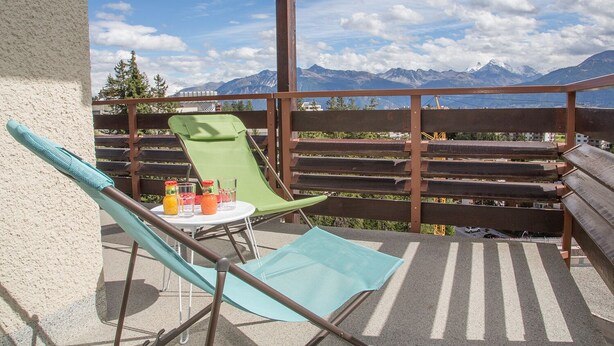 Balkon med udsigt til bjerge, en grøn stol, en blå stol og et lille bord med drikkevarer.