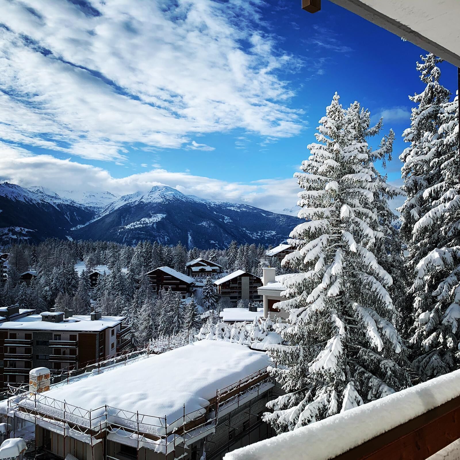 A snow-covered mountain landscape with a clear sky, a view of a ski resort with buildings, and snow-laden trees.