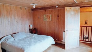 Una habitación con paneles de madera, una cama, una mesita con una lámpara y una puerta que da a otra habitación.