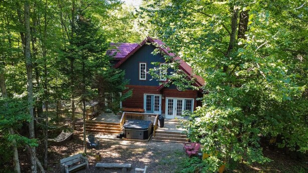 A cabin with a red roof, surrounded by dense forest. There is a wooden deck with a hot tub and a bench. - "Dingle Run Lodge" at Sir Sam's (hot tub/sauna) (EAGLE)