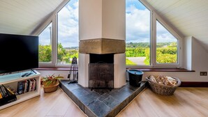 A modern living room with a stone fireplace, large windows, a bookshelf, and a television. - Peaceful countryside retreat with sea views (Penzance)