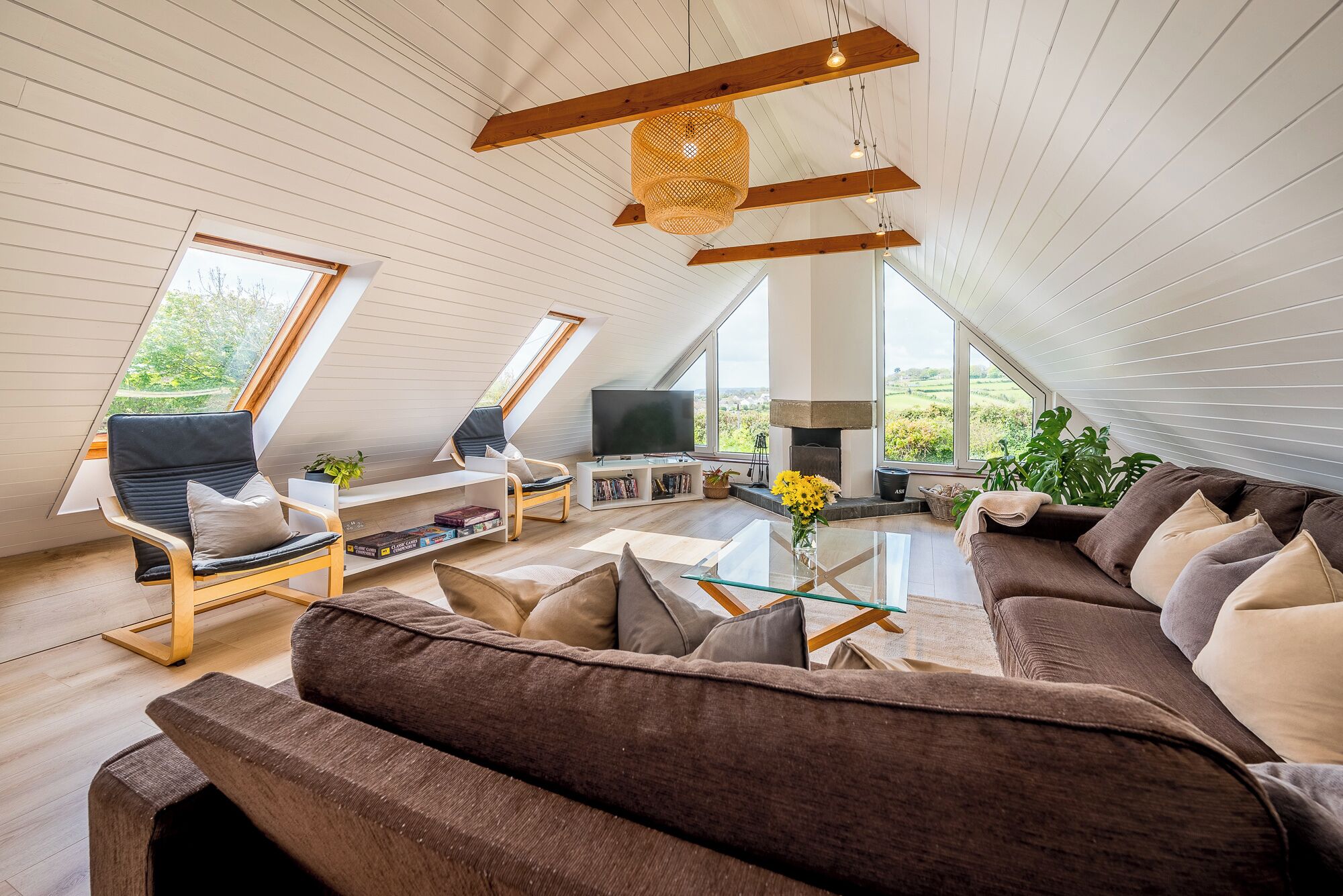 Un salon moderne de style industriel, avec un grand canapé d’angle, une table basse en verre et un téléviseur. Deux grandes fenêtres laissent entrer la lumière naturelle et offrent une vue sur l’extérieur.
