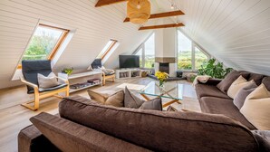 A modern loft-style living room with a large sectional sofa, a glass coffee table, and a TV. There are two high windows providing natural light and views of the outdoors. - Peaceful countryside retreat with sea views (Penzance)