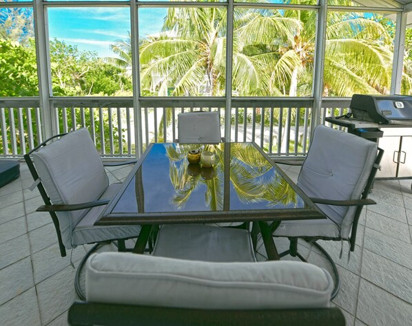 A glass-top table with two chairs, a small table lamp, and a grill on a balcony with a view of palm trees.
