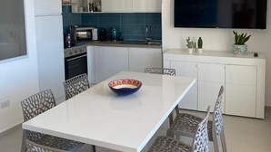 Una cuina moderna amb armaris blancs, un televisor gran i una zona de menjador amb taula blanca i cadires estampades.