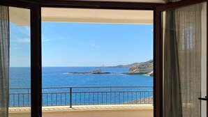 Una habitació amb un gran finestral corredís que dona a un balcó amb vista al mar i a la costa rocosa.