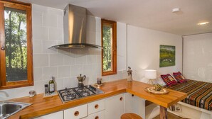 Eine kleine Küche mit Herd, Spüle und einer Holzauszugplatte. Zwei Hocker und ein Bett mit bunter Decke.